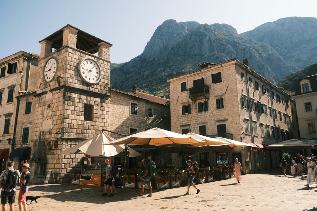 The leaning 1602 Clock Tower on the Square of Arms in Kotor's Old Town, with cafe umbrellas and mountains behind